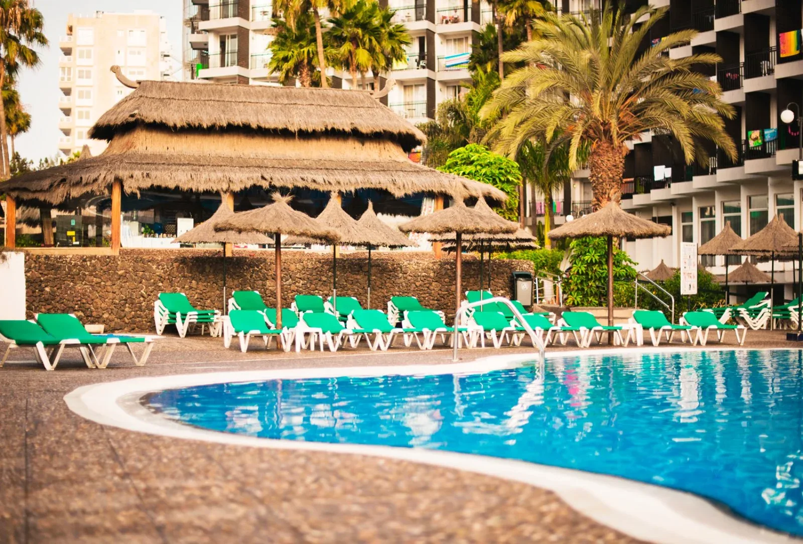Bordo piscina con lettini verdi, ombrelloni di paglia e edificio dell'hotel sullo sfondo